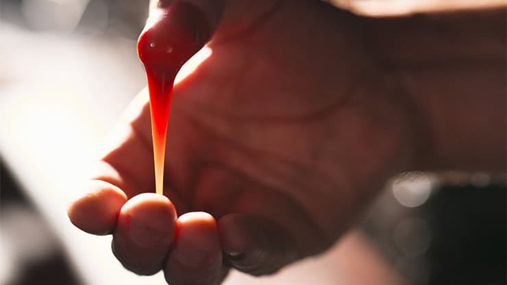 Close-up of a hand stretching a droplet of red liquid with a thin stream falling.