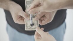 Close-up of a hand with black lipstick applying it on lips, with cash in the background.