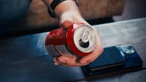 Up of a hand holding a soda can with a smartphone nearby.