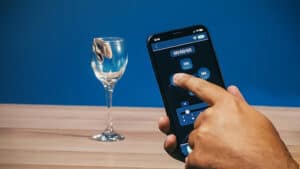 Close-up of a hand recording a magic trick with a smartphone and a glass on a wooden table.