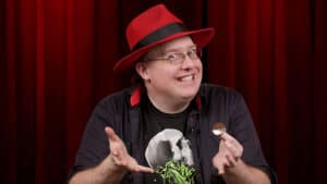 Magician Bizzaro performing a magic trick with a coin in front of a red curtain.