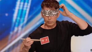 Cyril performing a card trick with a playing card and blindfold on stage.