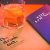 Close-up of a cocktail glass with an orange drink on a table with "The Drink Act" booklet.