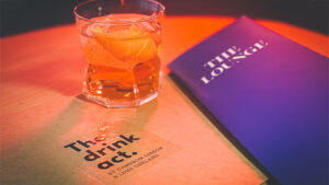 Close-up of a cocktail glass with an orange drink on a table with "The Drink Act" booklet.