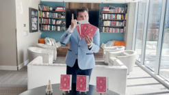Magician performs Jumbo McDonald Aces card trick with red-backed cards in a modern living room.