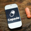 Close-up of a smartphone displaying "Capsule" with a pill-shaped icon on a wooden surface.