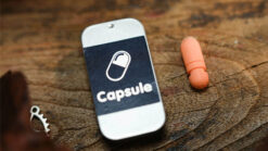 Close-up of a smartphone displaying "Capsule" with a pill-shaped icon on a wooden surface.